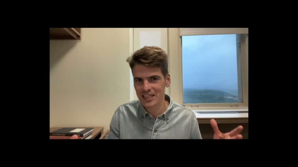 Person sitting in an office, gesturing with hands, with a window showing a cloudy sky in the background.