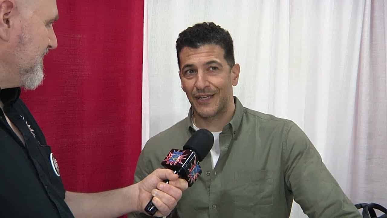 A man in a green shirt is being interviewed by another person holding a microphone with a colorful logo. They are indoors, standing in front of red and white curtains.