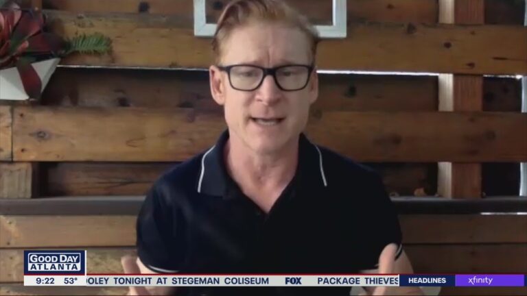 Man speaking during a video call on "Good Day Atlanta," with wooden wall background. News ticker visible at the bottom.