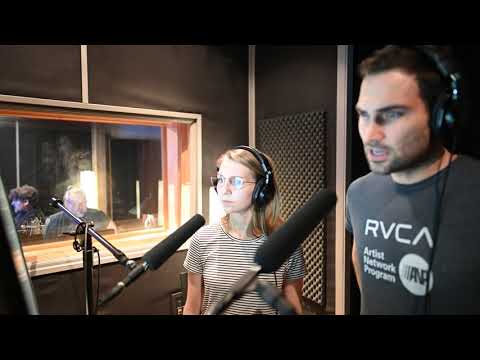 Two people wearing headphones stand in front of microphones in a recording studio. A man and a woman are visible through a window in the background.