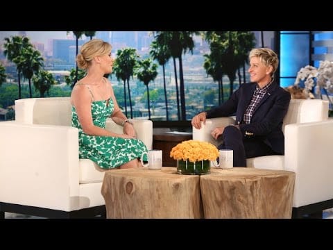 Two people sitting and talking on a talk show set, surrounded by a scenic backdrop with palm trees and a table with a flower arrangement between them.