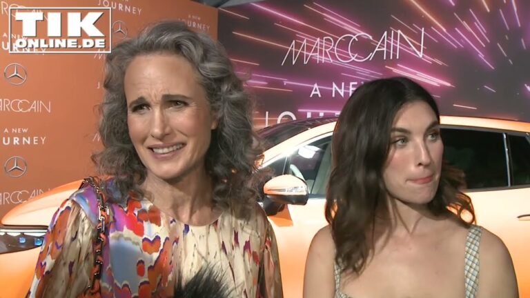 Two women stand in front of a backdrop with colorful lights and logos. One has curly gray hair and wears a patterned dress; the other has straight dark hair and wears a sleeveless dress.