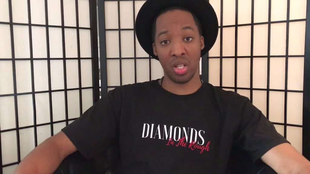 A person wearing a black hat and a "Diamonds in the Rough" T-shirt sits in front of a black and white folding screen.