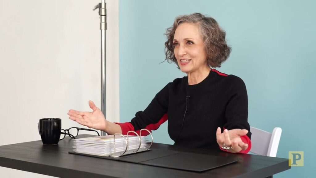 An older adult with short gray hair, wearing a black top, sits at a table with a binder and a mug, gesturing with both hands against a blue background.