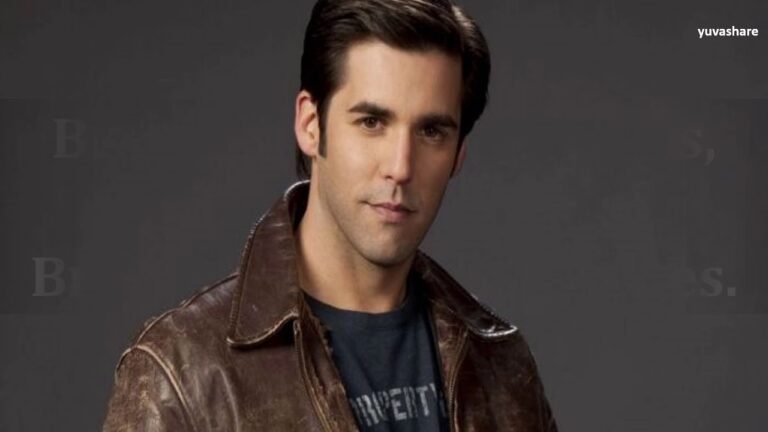 A person with dark hair wearing a brown leather jacket over a black shirt stands against a dark gray background.