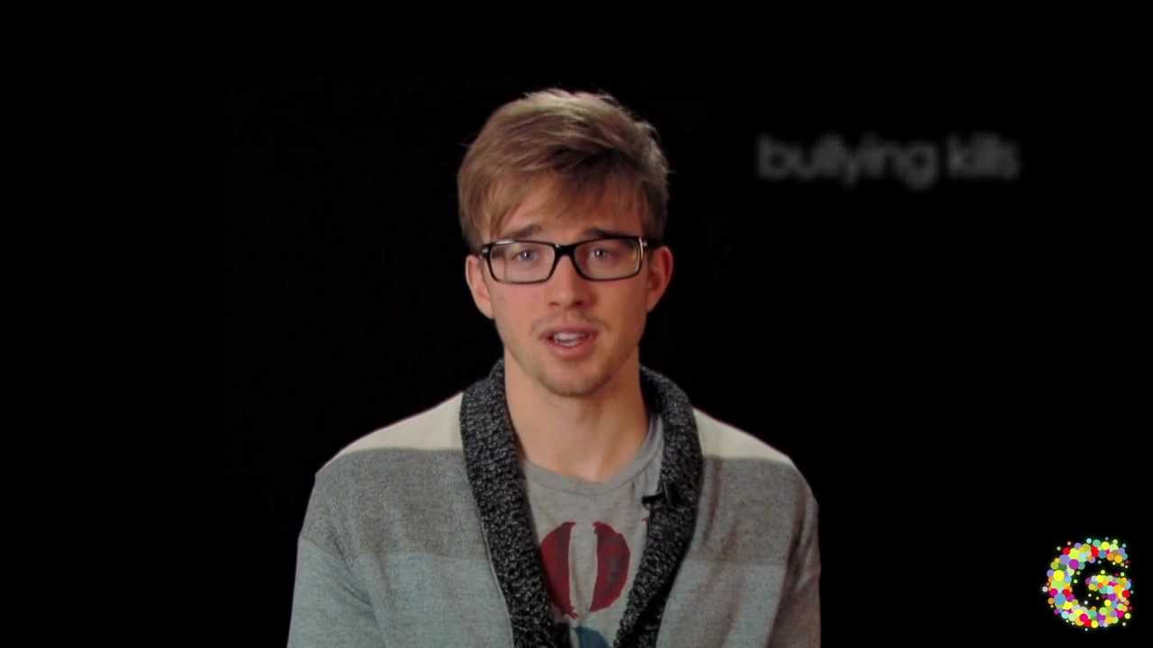 A person wearing glasses and a sweater speaks in front of a black background with the text "bullying kills" partially visible.