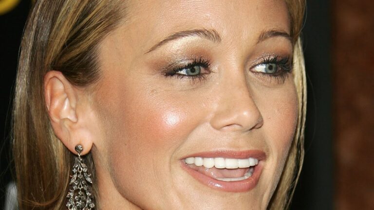 A close-up of a person with light brown hair, wearing metallic eyeshadow and long earrings, smiling against a dark background.