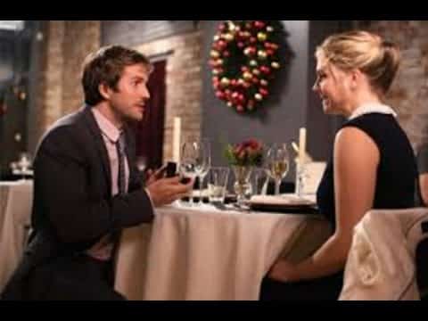 A man kneels and holds a ring box towards a woman seated at a restaurant table. A festive Christmas wreath is on the wall behind them.