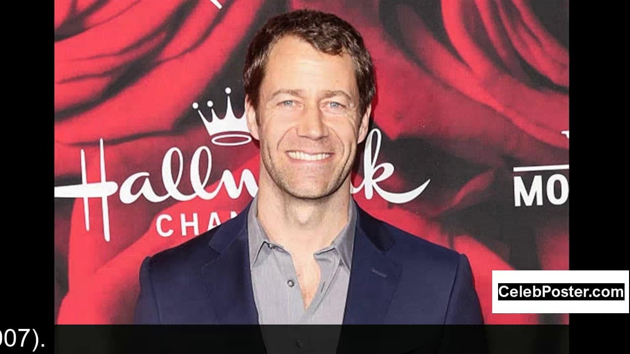 Smiling man in a dark suit stands in front of a Hallmark Channel backdrop with red flowers.