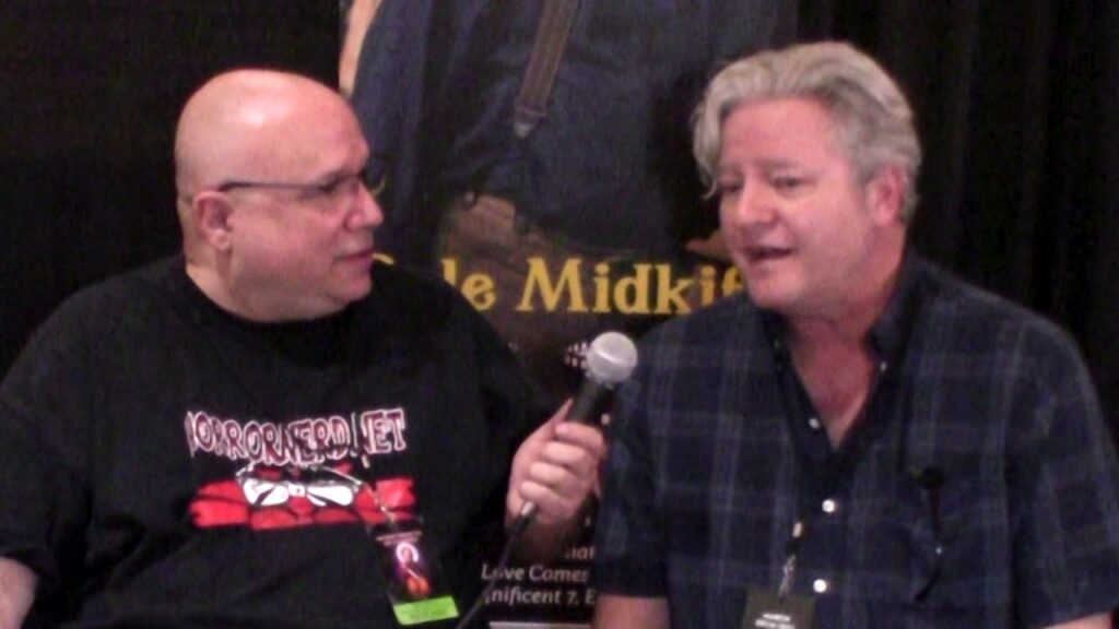 Two men seated, one holding a microphone, likely conducting an interview. A poster is visible in the background.