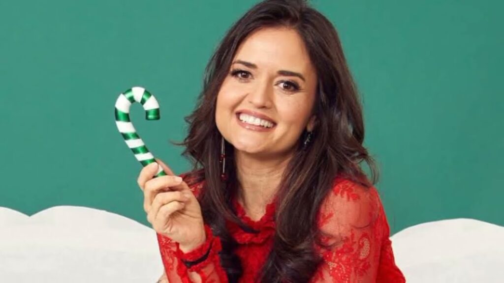 A woman in a red lace top smiles while holding a green and white candy cane against a green background.