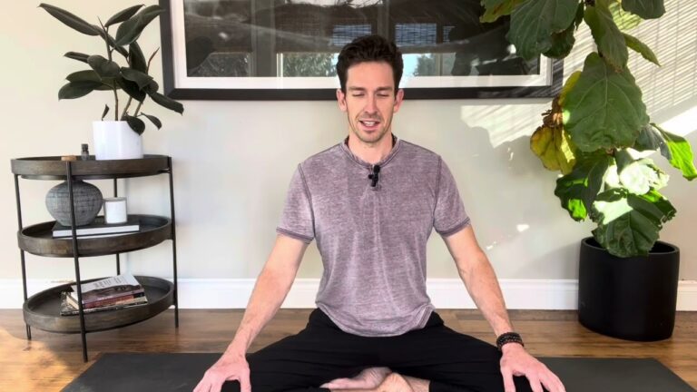 A person sits cross-legged on a yoga mat in a room with plants and a round shelf, with a large framed picture on the wall behind.