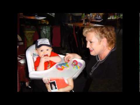 A baby in a high chair wearing a hat and red shirt holds a toy. A smiling woman is seated next to the baby.