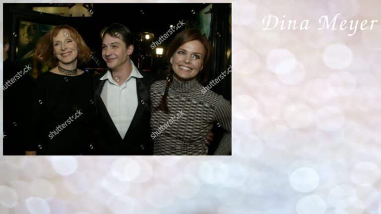 Three people posing together, two women and one man in a suit, smiling at an event. The background is blurred with soft, warm lighting.