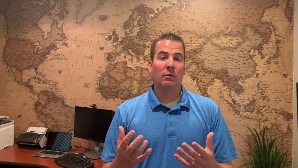 A person in a blue polo shirt is speaking in front of a desk and a map-covered wall.