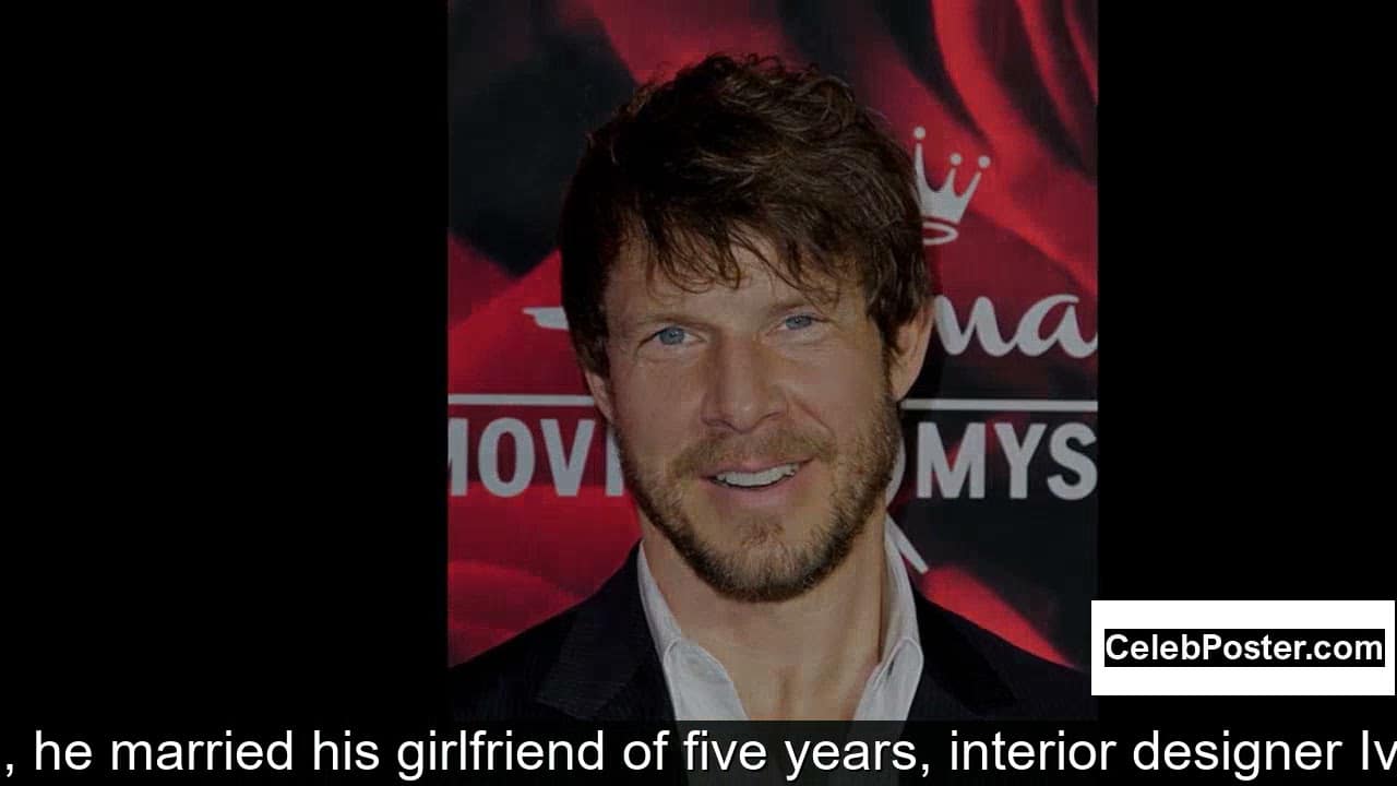 A man with brown hair and a beard, wearing a dark suit and light blue shirt, smiles in front of a red and black backdrop with text.