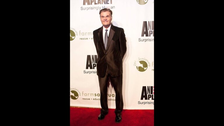 A man in a suit stands on a red carpet in front of a backdrop with logos and text.