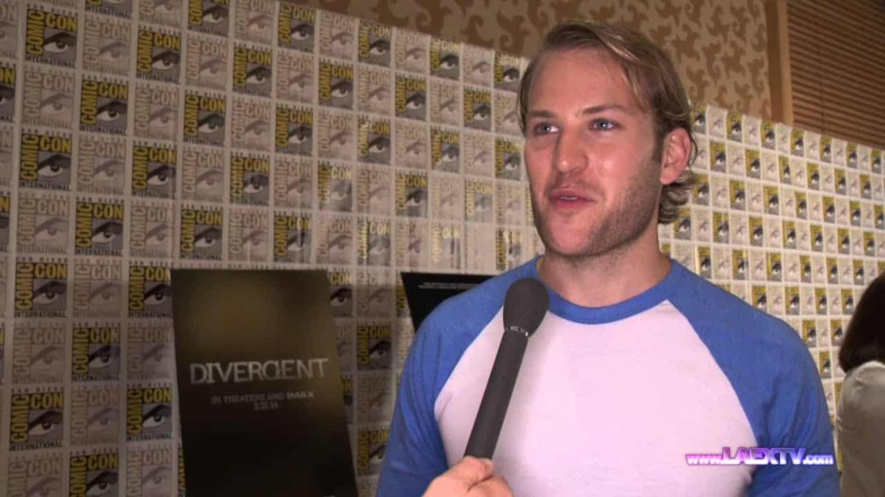 Person being interviewed at an event, standing in front of a wall covered with repeated logos and a "Divergent" poster.