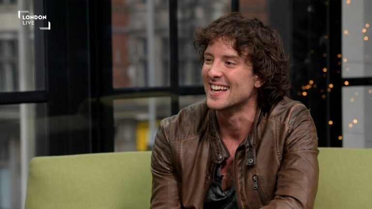 A person with curly brown hair is sitting on a sofa, wearing a brown leather jacket, smiling, with city buildings visible through the window behind them.