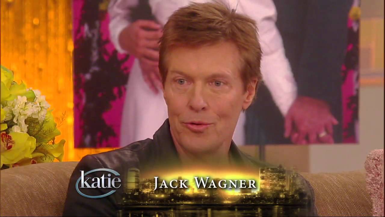 A person sitting on a talk show set with "Katie" and "Jack Wagner" displayed on the screen. A bouquet of flowers and a blurred photo are in the background.