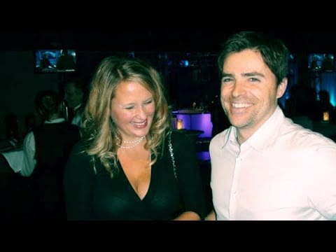 A woman and a man smile at a social gathering, with dim lighting and a bar in the background.