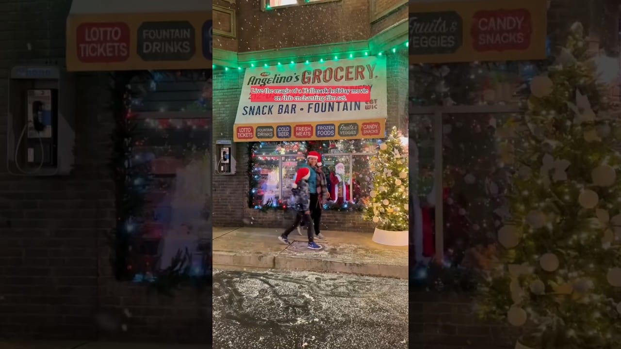 Two people walk past Angelino's Grocery, a store with a festive Christmas window display and lights.