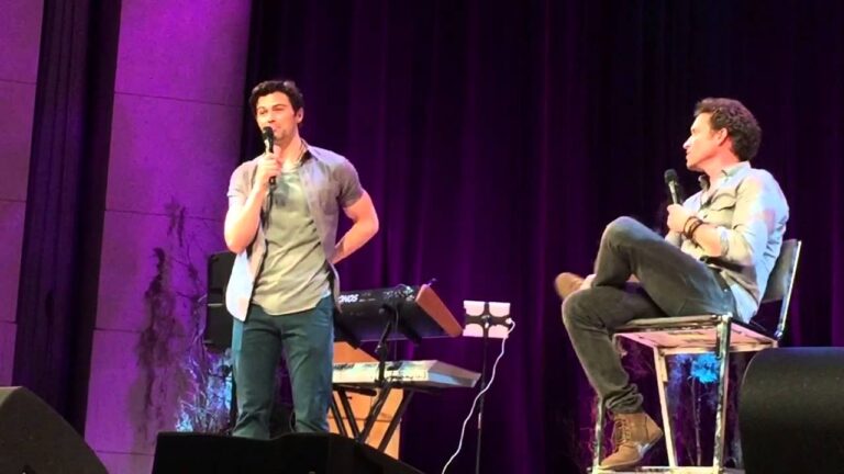 Two men on stage with microphones; one sitting on a chair, the other standing near a keyboard. The backdrop is purple-lit.