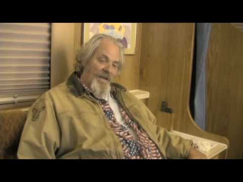 An older man with gray hair and a beard, wearing a beige jacket over an American flag shirt, sitting indoors near a window and wood-paneled wall.