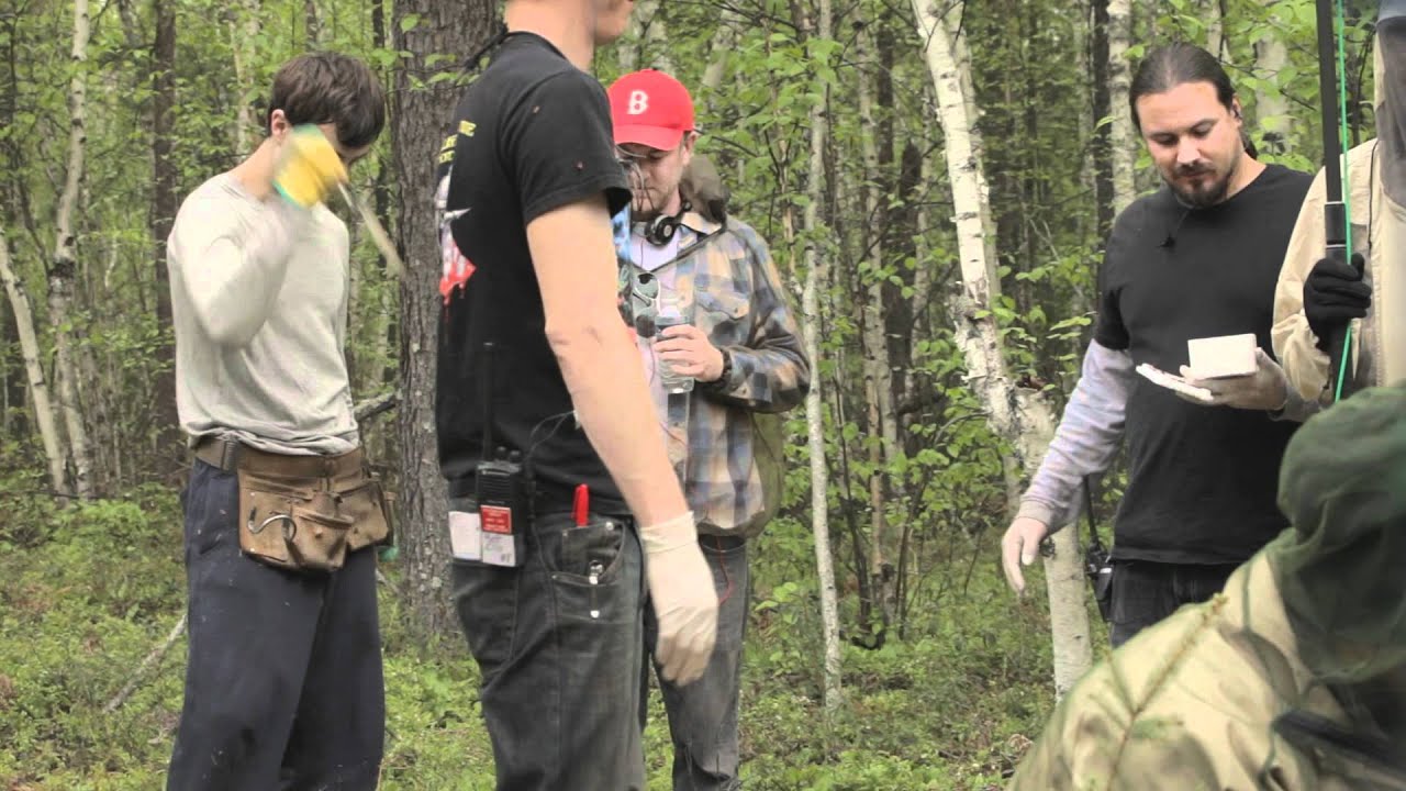 Four men are standing in a forest, some wearing gloves and holding tools. One man is looking at a notepad. Dense trees are in the background.