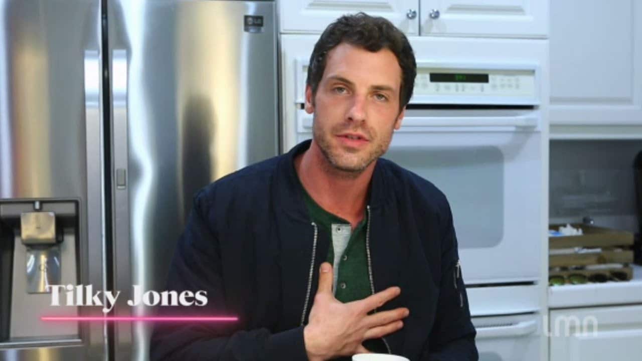 A man in a kitchen stands in front of a refrigerator and microwave, with a hand on his chest, wearing a dark jacket.