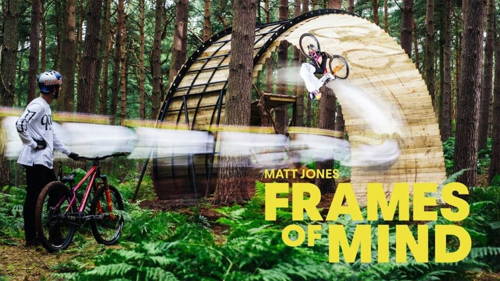 A mountain biker rides up a wooden ramp in a forest as another cyclist watches. Text reads, "Matt Jones: Frames of Mind.