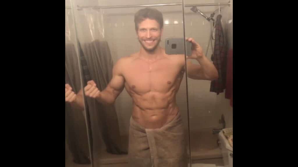 A person takes a mirror selfie in a bathroom, showing a muscular physique, with a towel wrapped around the waist.