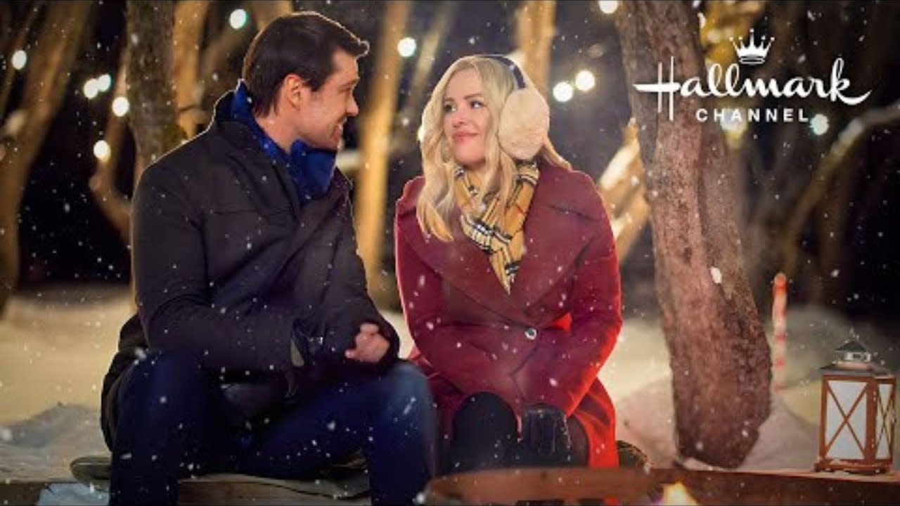 A man and woman sit together in a snowy outdoor setting, both dressed warmly. The woman wears earmuffs and a red coat. Hallmark Channel logo is in the top right corner.