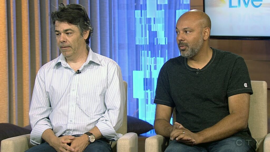 Two men seated indoors, one in a striped shirt and the other in a dark t-shirt, with a blurred backdrop featuring curtains and a lit screen.