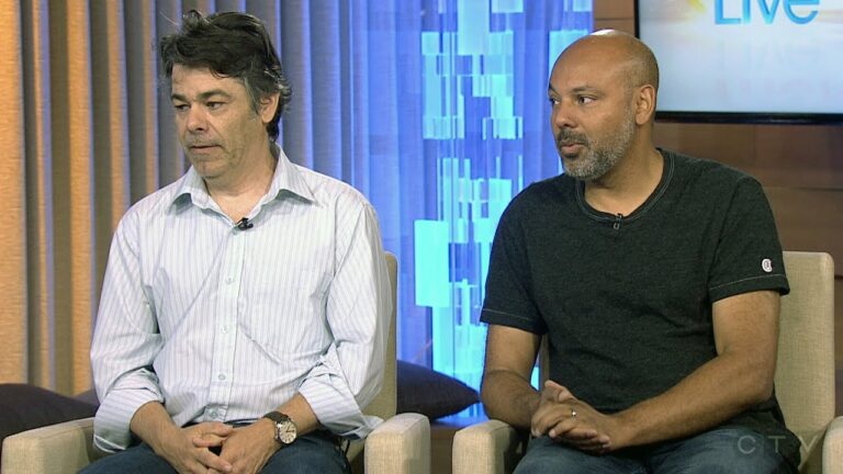 Two men seated indoors, one in a striped shirt and the other in a dark t-shirt, with a blurred backdrop featuring curtains and a lit screen.