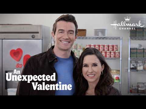 A man and a woman smiling, standing in a kitchen decorated with heart-shaped ornaments. Text reads "Hallmark Channel: An Unexpected Valentine.