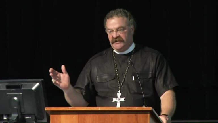 A man in clerical attire speaks at a podium, gesturing with one hand. A computer monitor is visible to his left.