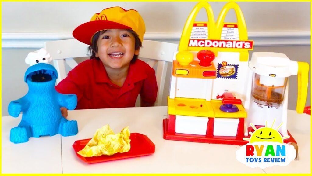 A child in a red outfit and yellow hat sits at a table with a McDonald's playset and a blue toy character.