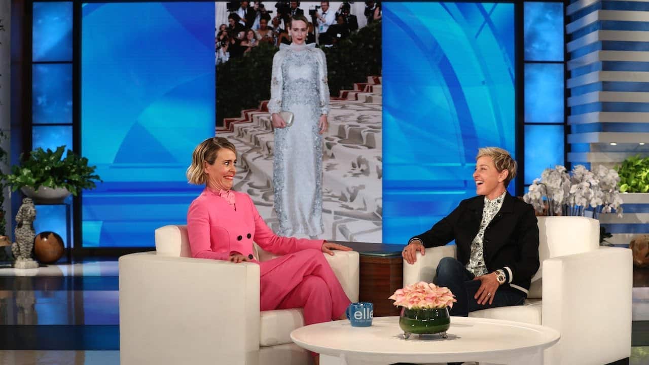 Two people are sitting in armchairs during a talk show. A large screen behind them displays a person in a silver gown.