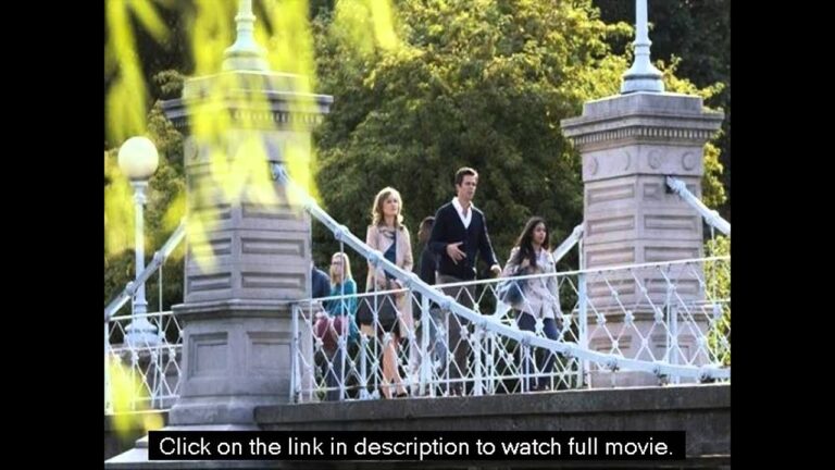 Four people walking across a decorative suspension bridge in a park setting with trees in the background.