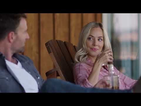 A man and a woman sit on wooden chairs. The woman is holding a bottle and looking at the man.