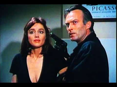 A man holds a gun to a woman's head in a tense indoor setting. A Picasso poster is partially visible in the background.