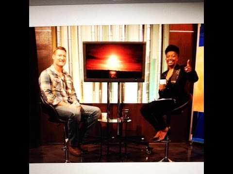 Two people are seated on a talk show set with a TV displaying a sunset in the background. One person is giving a thumbs up.