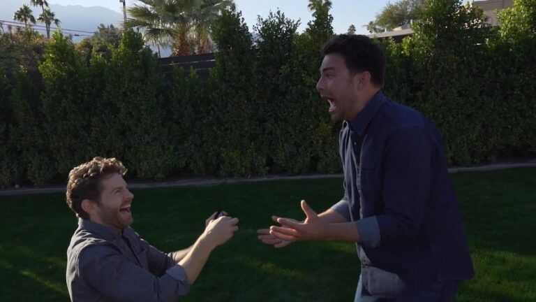 One person kneels on the grass holding a small box, while the other person reacts with excitement. Trees and mountains are in the background.