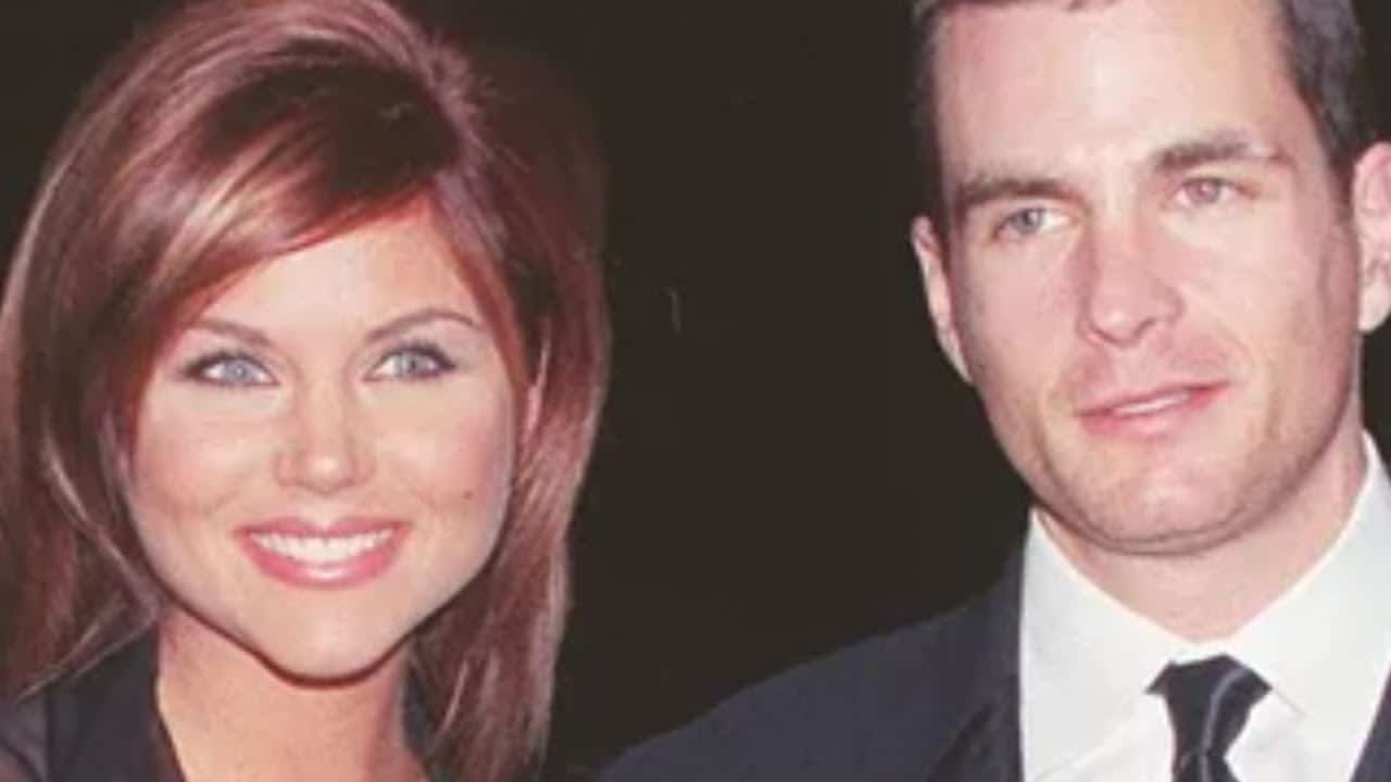 A man and a woman smiling, both wearing formal attire, with the woman in a dark dress and the man in a suit and tie, standing against a dark background.