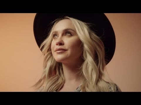 A person with long blonde hair wearing a black hat looks slightly upwards against a soft orange background.