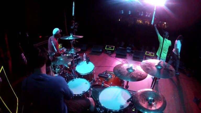 View from behind a drummer on stage, showing a band performing under colorful lights to an audience.