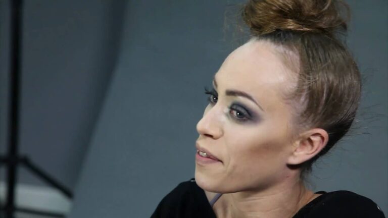 Person with hair in a bun, wearing makeup, and looking to the side against a neutral background.