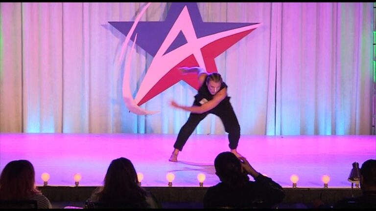 Person performs a ribbon dance on stage with a large star logo in the background, while spectators watch from the audience.