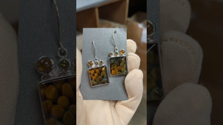 A pair of dangle earrings featuring square pendants with a yellow and black circular pattern, held by a gloved hand.